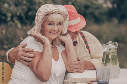 Happy relationship of an old couple. Smiling little old grandparents are sitting at the table and talking about the memories of youth. Drink lemonade with fruit in the garden