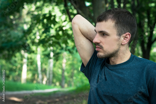 Portrait of a pensive young man in a dark green T-shirt, serious emotions, in the background a park or a forest, trees and mustaches everywhere, a hand raised up and bent at the elbow, a deep look