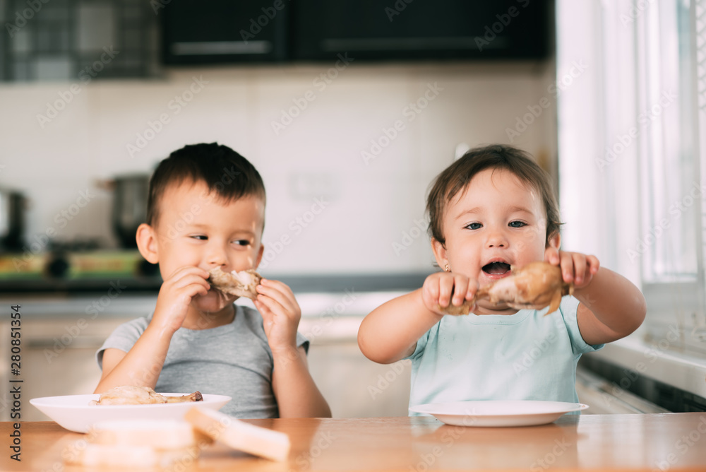 Kids boy and girl , brother and sister in the kitchen having fun and eating chicken with appetite - obrazy, fototapety, plakaty 