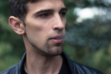 dramatic portrait of a young, brutally-looking young man who smokes a cigarette smoke in the woods. Close-up. Fashionable hairstyle and manly bristles. Wearing a black leather jacket.