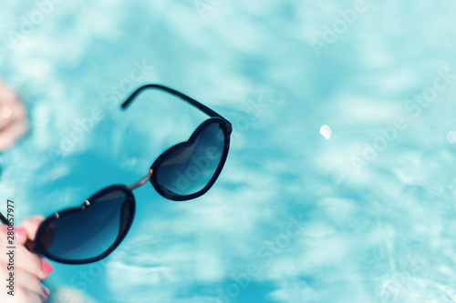 Women's hand with a beautiful manicure holding sunglasses in shape of heart on a background against the azure water of the pool. The concept of summer, love and relaxation. Fashion summer accessory