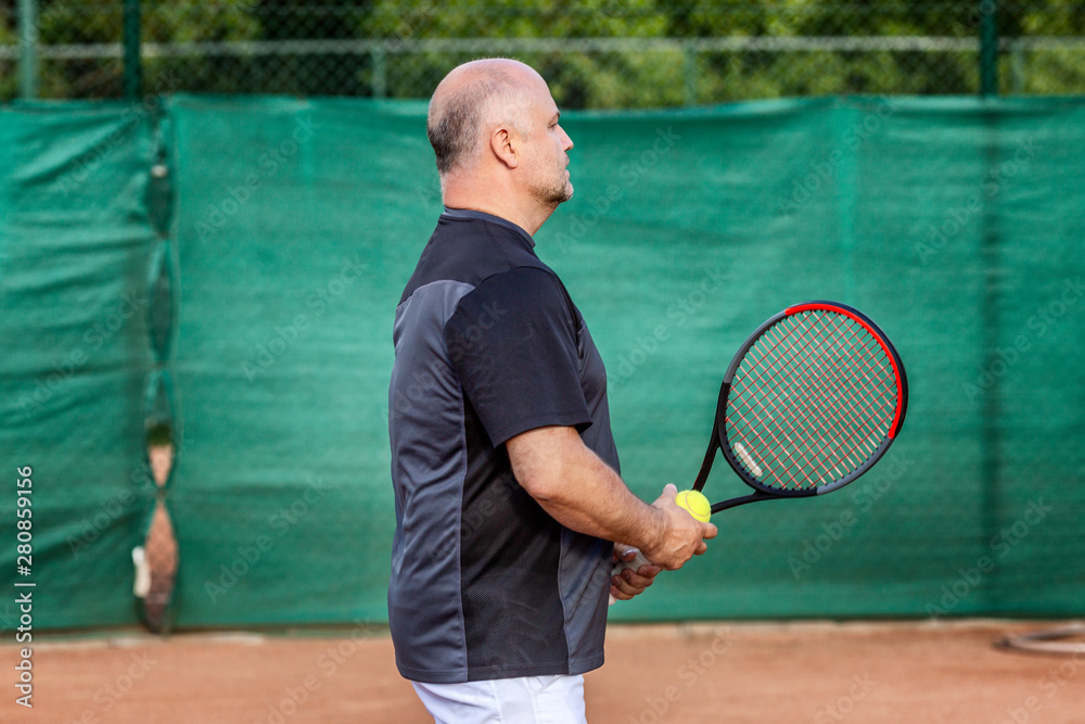 Adult bald man plays tennis on the court. Sunny day.