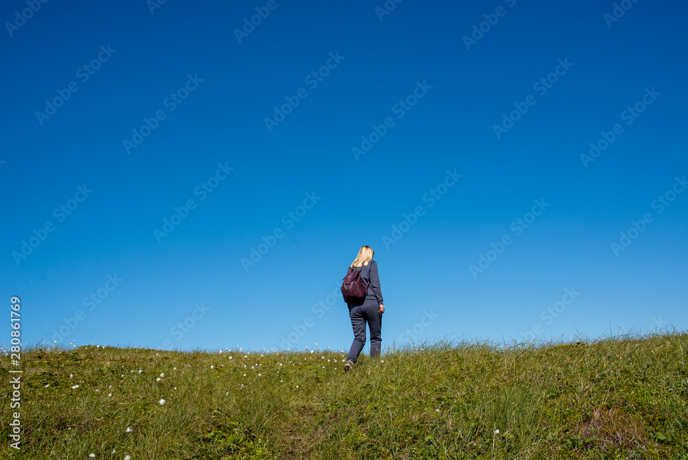 A girl with backpack outdoors rises up. Green grass and blue sky. Leisure activity, hiking. Enjoy the moment, relax. Wanderlust. Adventure, freedom, lifestyle. Explore North Norway. Summer Scandinavia