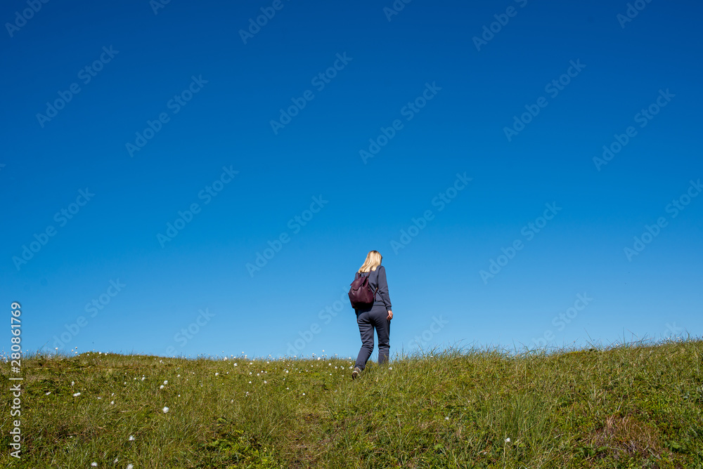 A girl with backpack outdoors rises up. Green grass and blue sky. Leisure activity, hiking. Enjoy the moment, relax. Wanderlust. Adventure, freedom, lifestyle. Explore North Norway. Summer Scandinavia A girl with backpack outdoors rises up. Green grass and blue sky. Leisure activity, hiking. Enjoy the moment, relax. Wanderlust. Adventure, freedom, lifestyle. Explore North Norway. Summer Scandinavia