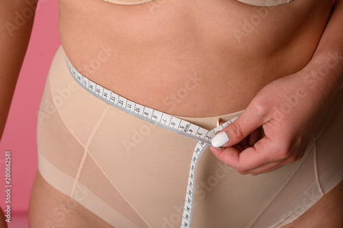 young woman size plus measuring her waist with a tape measure on pink background