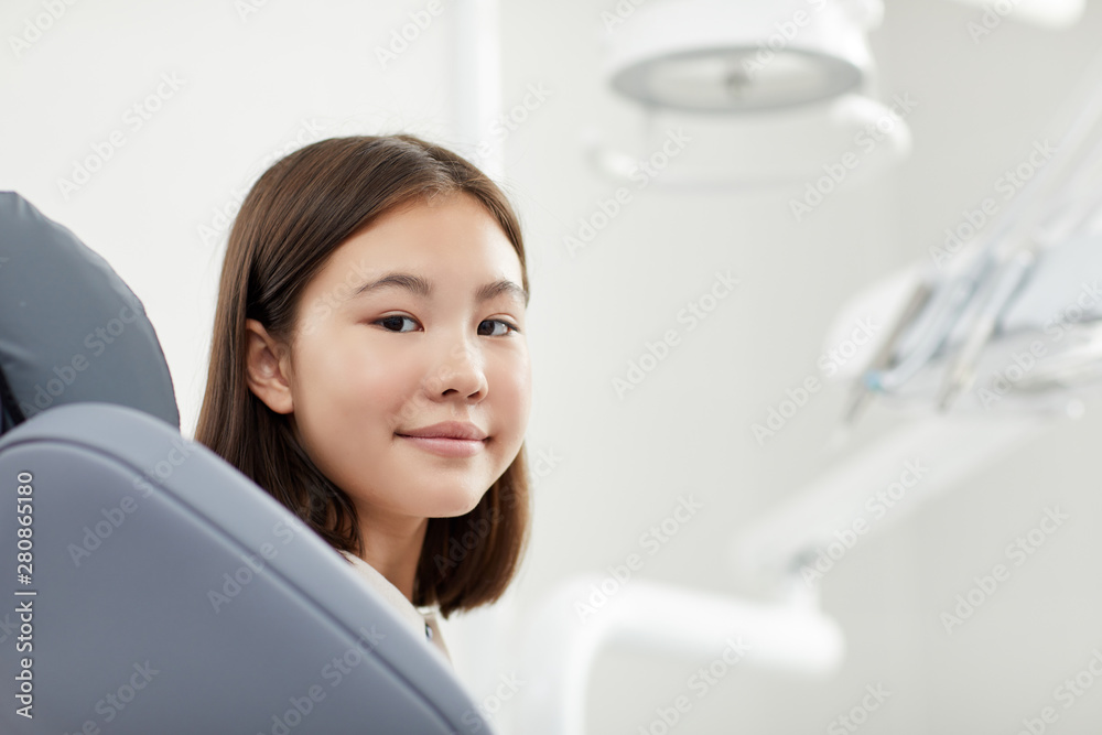 Portrait of teenage Asian girl sitting in dental chair in modern clinic and smiling at camera , copy space Portrait of teenage Asian girl sitting in dental chair in modern clinic and smiling at camera , copy space