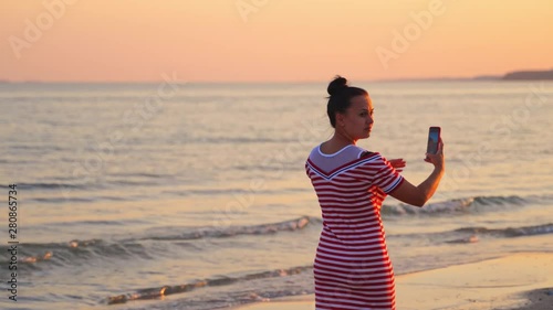 Woman takes picture of beautiful sunset sea with phone. Girl in dress holds smartphone making photo memories of summer vacation with mobile camera. photo