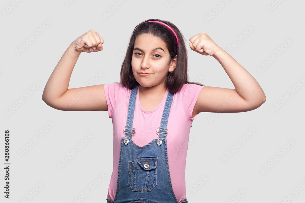 I am strong. Portrait of satisfied brunette young girl in casual pink t-shirt and blue overalls standing with raised arms and looking at camera. indoor studio shot, isolated on light gray background. I am strong. Portrait of satisfied brunette young girl in casual pink t-shirt and blue overalls standing with raised arms and looking at camera. indoor studio shot, isolated on light gray background.