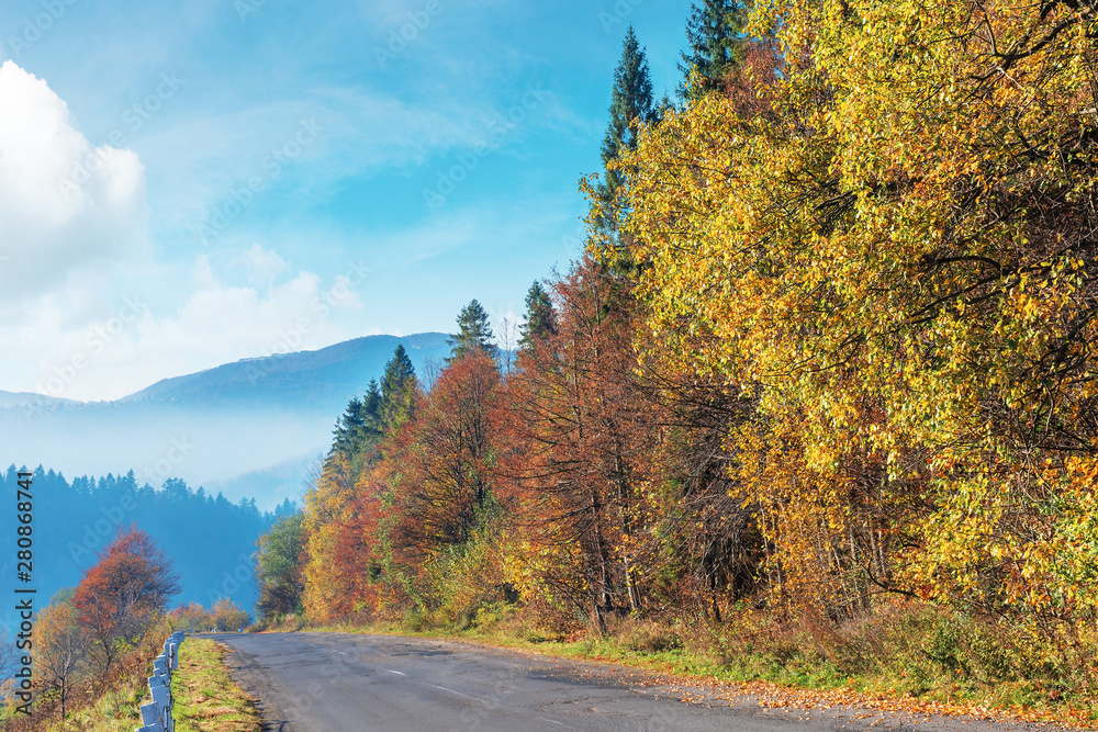 Fototapeta premium old cracked asphalt road in mountains. straight path along the forest on hill. trees in fall colors. distant ridge in haze. sunny autumn weather with fluffy clouds on the bright blue sky