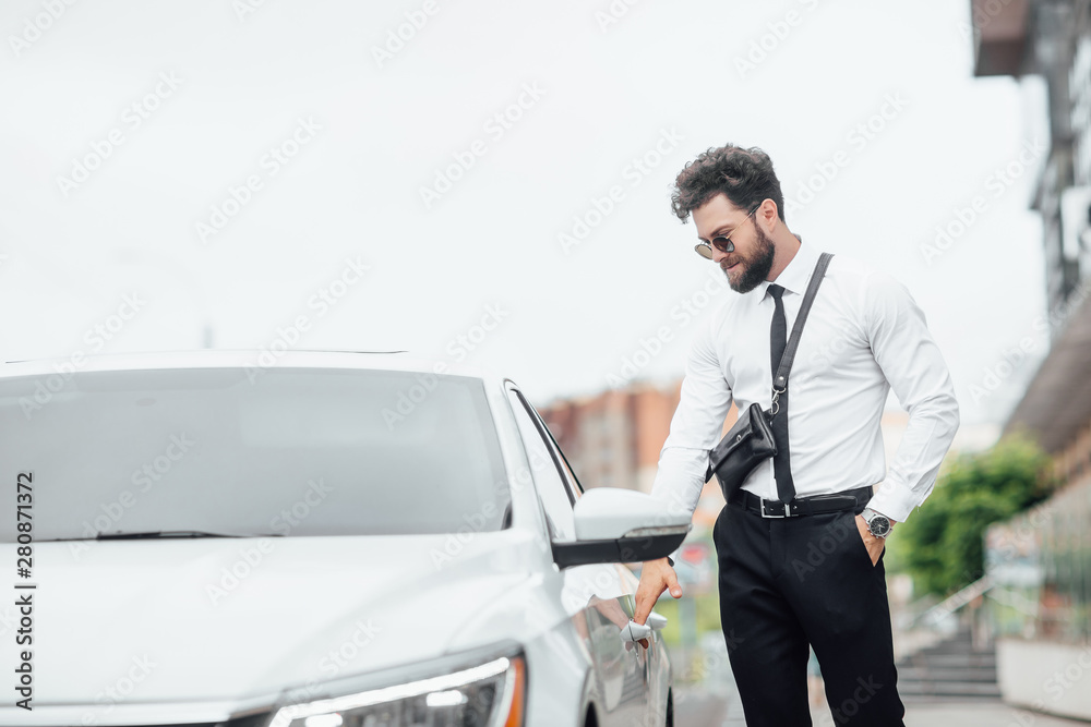 A handsome successful businessman in business clothes opens his car on the streets of the city near the modern office center. Luxury life. A handsome successful businessman in business clothes opens his car on the streets of the city near the modern office center. Luxury life.