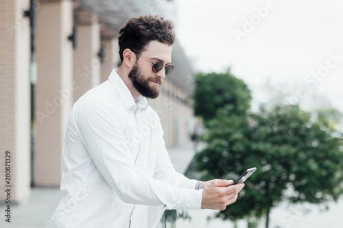 A bearded, serious, stylish man in white shirt and sunglasses standing on the streets of the city and surfing smartphone.