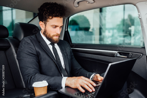 Handsome bearded manager working on his laptop with coffee to go on the backseat of the new car.