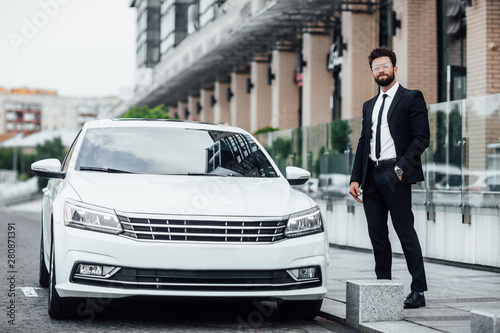 Young successful businessman in black suit on the streets of the city with a new white car near a modern business center. © Тарас Нагирняк