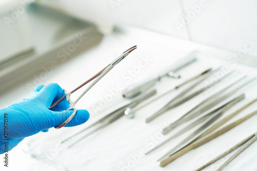 Hand of a nurse in a blue medical glove is holding the clip. Medical instruments for surgery are on metal table in the operating room in the background.