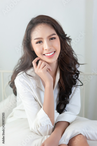 A beautiful Asian woman in a sexy white shirt smiling happily on a white bed.