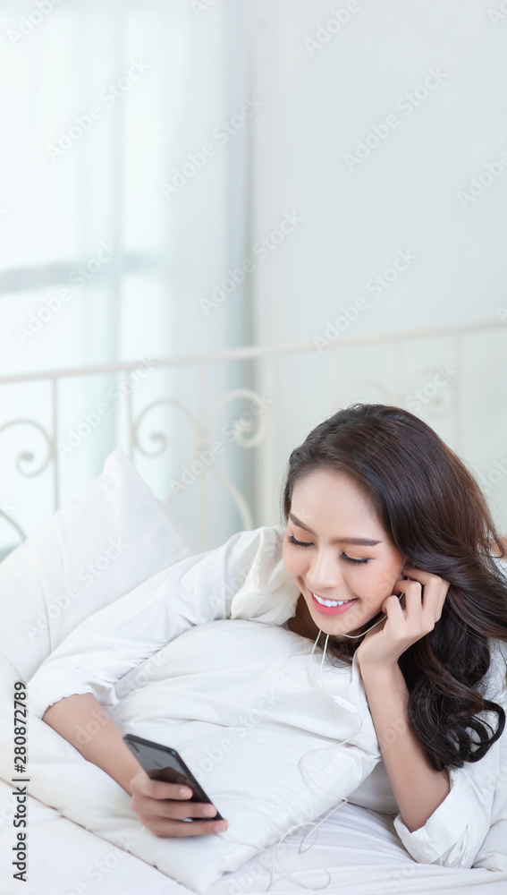 A beautiful Asian woman in a white shirt listening to music and smiling happily on a white bed. A beautiful Asian woman in a white shirt listening to music and smiling happily on a white bed.