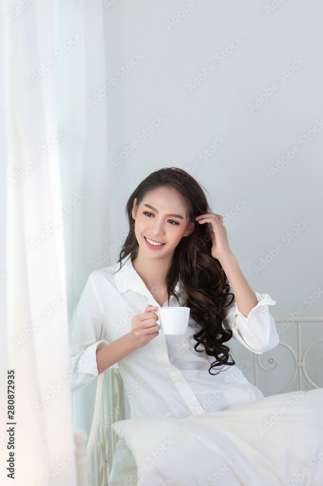A beautiful asian woman in a white shirt and coffee cup that is smiling happily in the white bed. A beautiful asian woman in a white shirt and coffee cup that is smiling happily in the white bed.