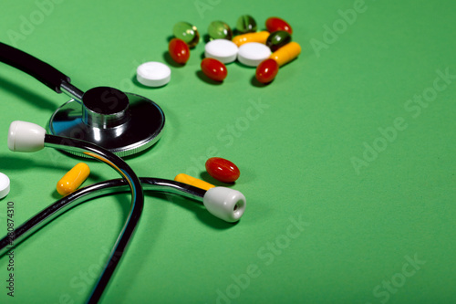 A group of bright medicine pills and a phonendoscope on a green background. Cropped shot, horizontal, side view. The concept of medicine and health. Crisis opioid painkiller and drug addiction concept
