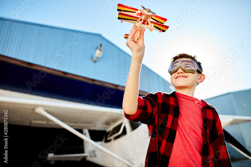 view of lovely kid playing with toy plane outdoor near the shed, white single-engine aircraft stands behind him, wears aviator glasses, red shirt and jeans. Aeromodelling hobby. Outdoor shot. photo