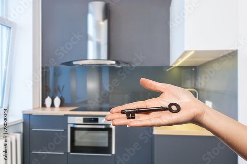 Business woman's hand holding key on blurry kitchen interior background. Real estate concept. Property For Sale. European furniture. Kitchen glass back wall. Modern kitchen design.