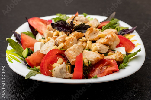 Warm salad with chicken, tomatoes, soft brynza cheese and greens, decorated with pine nuts. dressing with olive oil. White cup on a black table