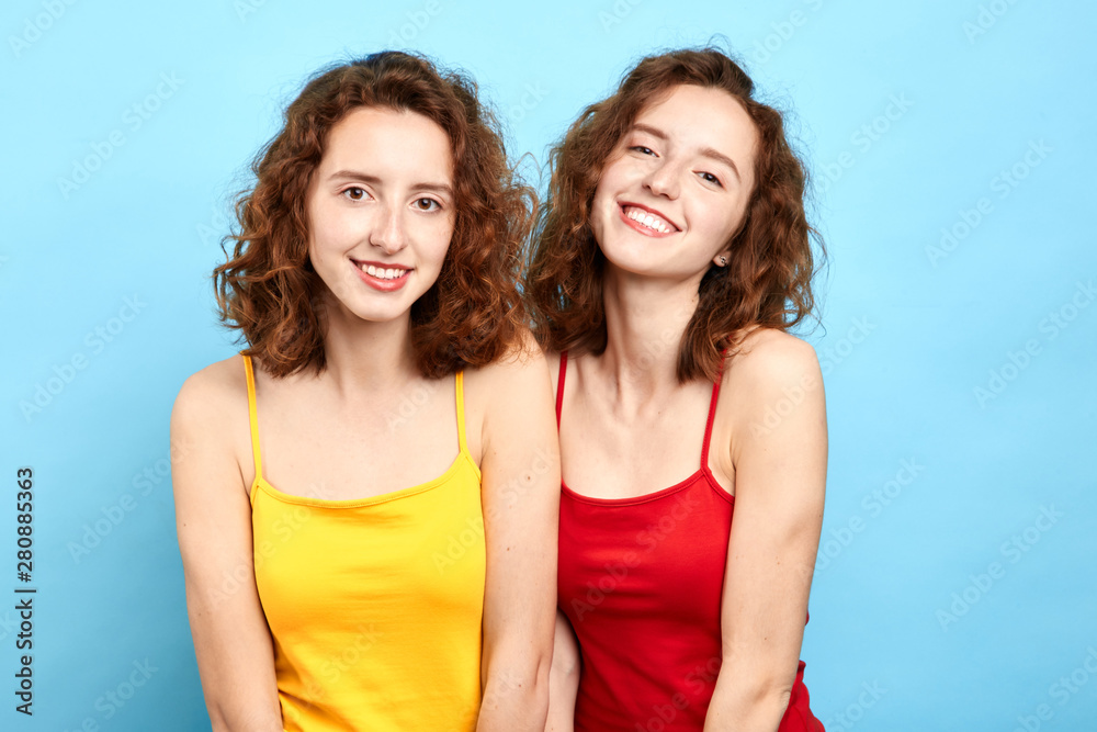 smiling twin sisters posing to the camera isolated on blue, beauty concept, girl in red T-shirt looks like the other one in yellow T-shirt. smiling twin sisters posing to the camera isolated on blue, beauty concept, girl in red T-shirt looks like the other one in yellow T-shirt.