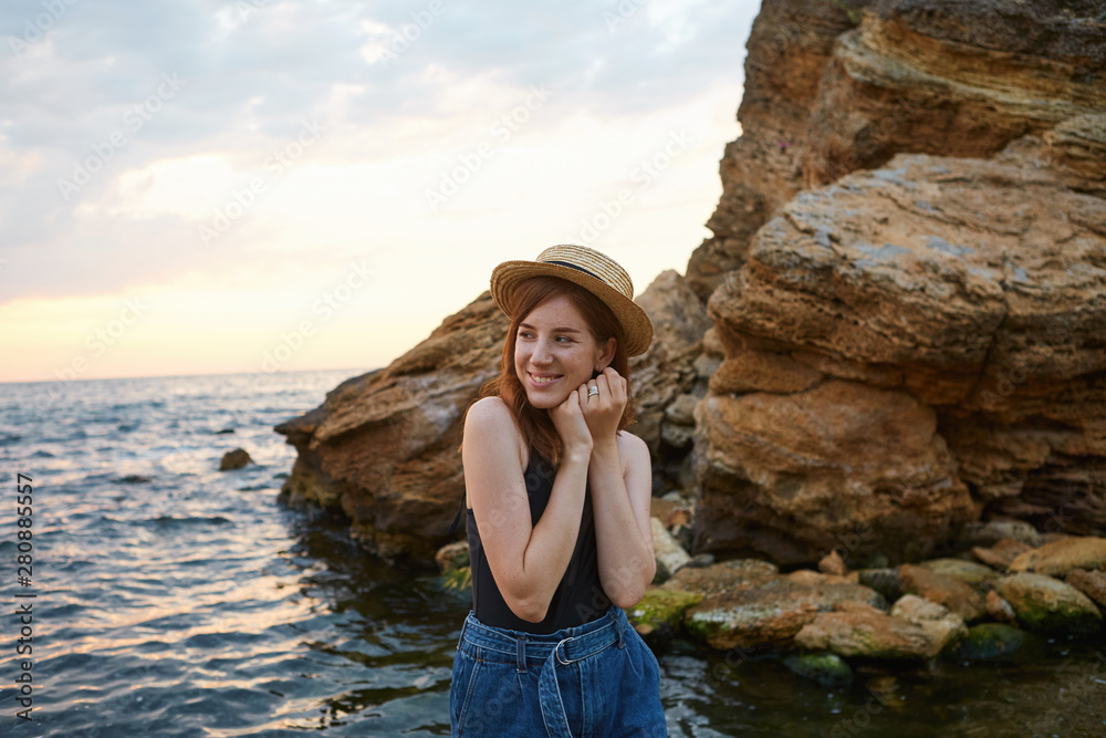 Young cute positive ginger girl with freckles wears in in hat, broadly smiles and enjoy the morning on the seaside, looks cheerful and happy.