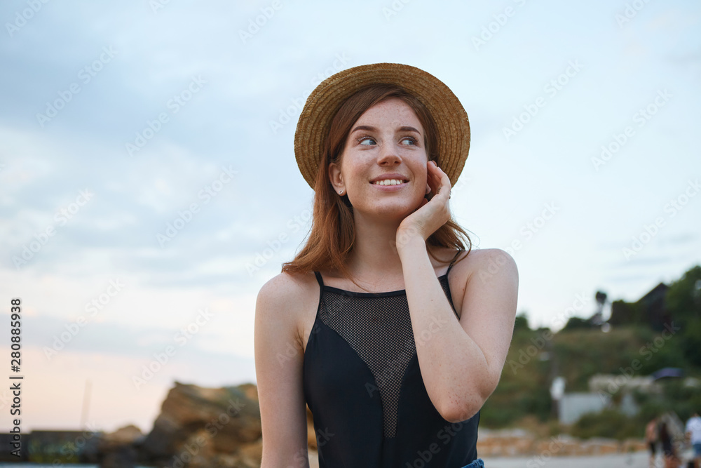 Portrait of young ginger cute freckles woman walks on the beach, wears hat, dreamily looks away and touches cheek, looks positive and happy.