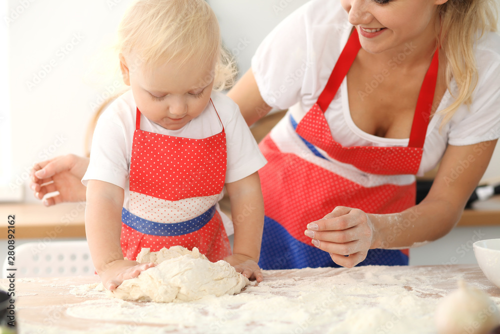 Little girl and her blonde mom in red aprons playing and laughing while kneading the dough in kitchen. Homemade pastry for bread, pizza or bake cookies. Family fun and cooking concept Little girl and her blonde mom in red aprons playing and laughing while kneading the dough in kitchen. Homemade pastry for bread, pizza or bake cookies. Family fun and cooking concept