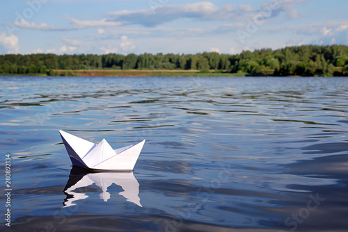 Small paper boat floating on blue calm river water, green forest, blue sky with light summer clouds on the horizon. Freedom, avdenture, dream and travel concept.