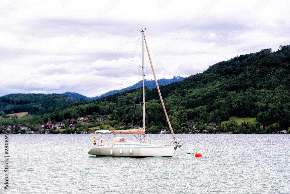 A white yacht in calm Kammersee's water. A cloudy day at Attersee lake at Salzkammergut area in Austria. There are a lot of green trees in the Alps mountains. The sky is full of grey clouds. A white yacht in calm Kammersee's water. A cloudy day at Attersee lake at Salzkammergut area in Austria. There are a lot of green trees in the Alps mountains. The sky is full of grey clouds.