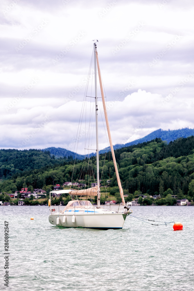 Fototapeta premium A white yacht in calm Kammersee's water. A cloudy day at Attersee lake at Salzkammergut area in Austria. There are a lot of green trees in the Alps mountains. The sky is full of grey clouds.