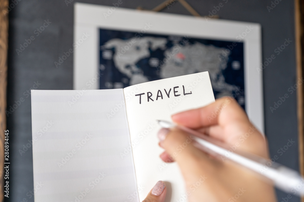 Female hands holding notebook and pen, making notes when planning a travel route and expenses in front of the map of the world. Close-up Female hands holding notebook and pen, making notes when planning a travel route and expenses in front of the map of the world. Close-up