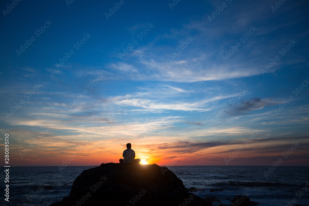 Silhouette of a lonely man sitting on the rocks near the ocean beach during the amazing sunset.