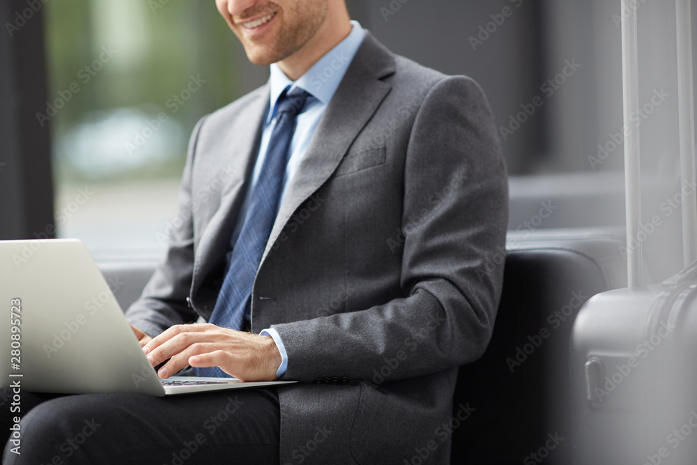 Close-up of optimistic young entrepreneur in suit sitting on sofa in airport and composing email for business partner on laptop