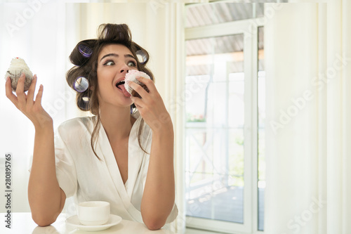 beautiful funny young brunette woman in hair curlers licking one cupcake and holding another in her hand sitting with cup of coffee in bright kitchen