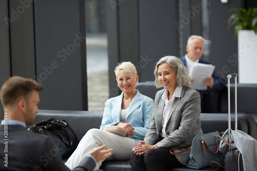 Cheerful mature business colleagues in jackets sitting on sofa in waiting area of airport and chatting while waiting for boarding