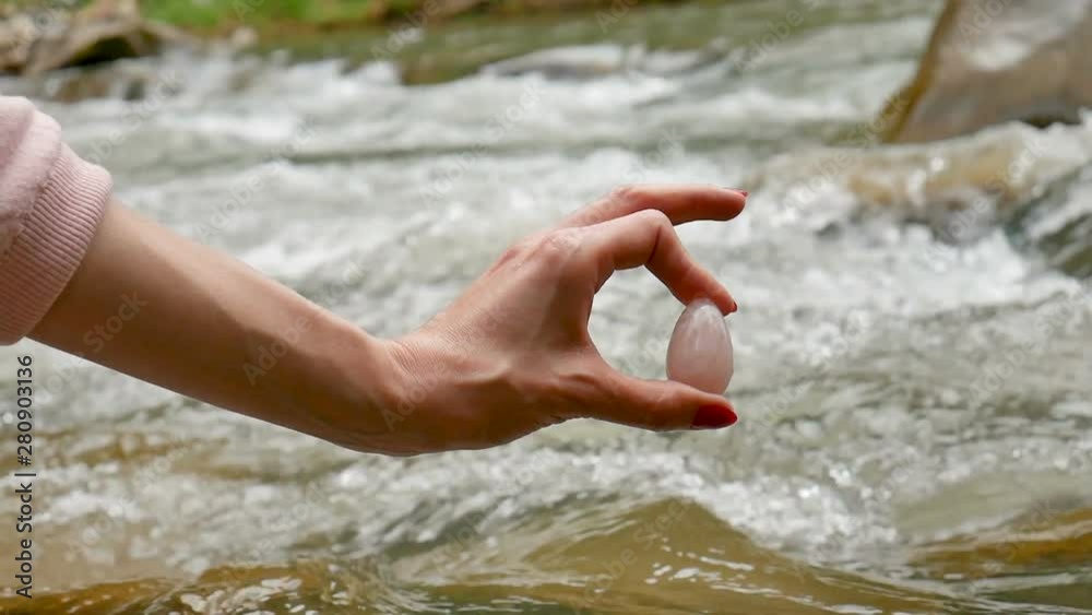 Female hand holding a rose quartz crystal yoni egg on river background. Women's health, unity with nature concepts. Female hand holding a rose quartz crystal yoni egg on river background. Women's health, unity with nature concepts.