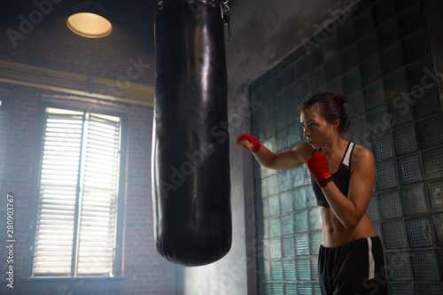 Dramatic portrait of tough female fighter hitting punching bag at boxing practice, copy space © Seventyfour