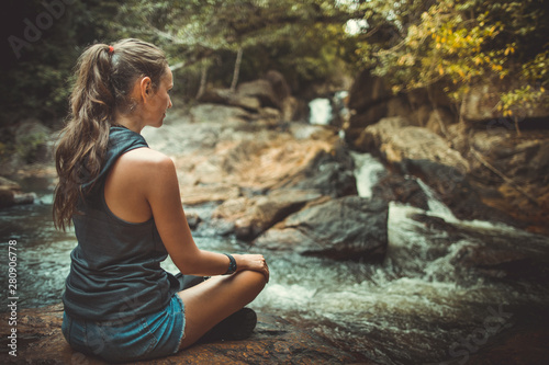 Young woman in yoga pose sitting near waterfall, Rear view