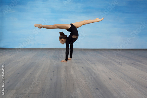 Girl gymnast in a black gymnastic swimsuit makes handstand legs divorced in splits on a blue sky background.