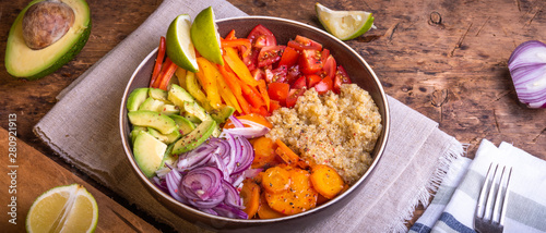 Bowl of salad with quinoa, carrots, avocado, onion, pepper, tomato with lime slices on a linen napkin on a wooden rustic table, top view