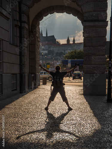 Fun in Prague - silhouette of a man posing beneath an arcade. He is turning his back to camera and throwing his shadow on a sidewalk in a stark back light. Sun lit Prague castle in distance.