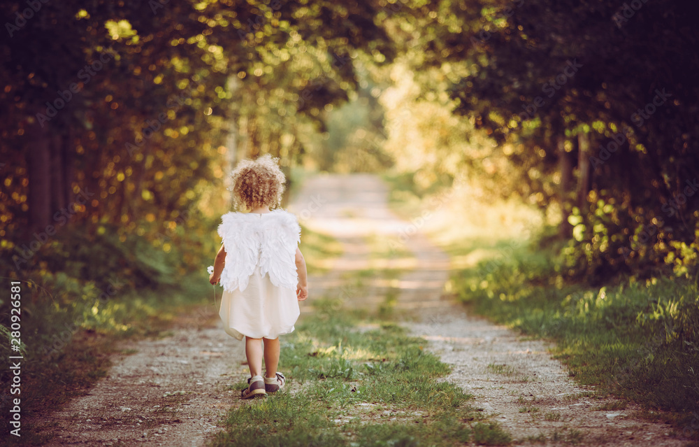 Super cute anonymous blonde curly hair girl child wearing white dress and angel wings, walking on countryside road. Backside view. Concept of child angel. Super cute anonymous blonde curly hair girl child wearing white dress and angel wings, walking on countryside road. Backside view. Concept of child angel.