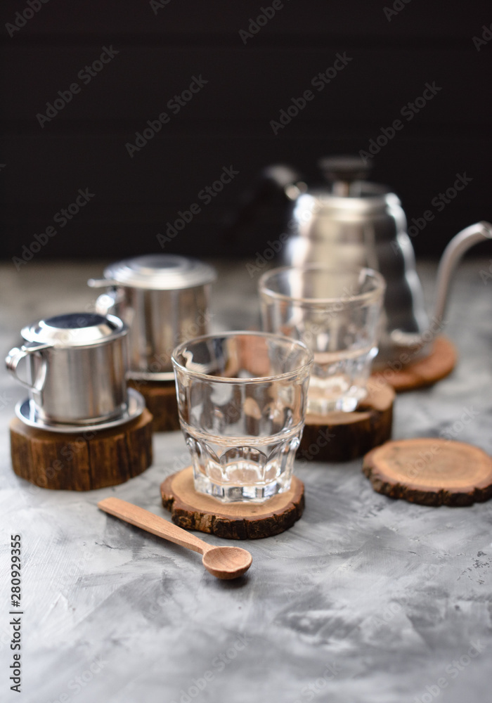 Equipment for making drip coffee Vietnamese style: phin, goose neck kettle and glasses on dark background Equipment for making drip coffee Vietnamese style: phin, goose neck kettle and glasses on dark background