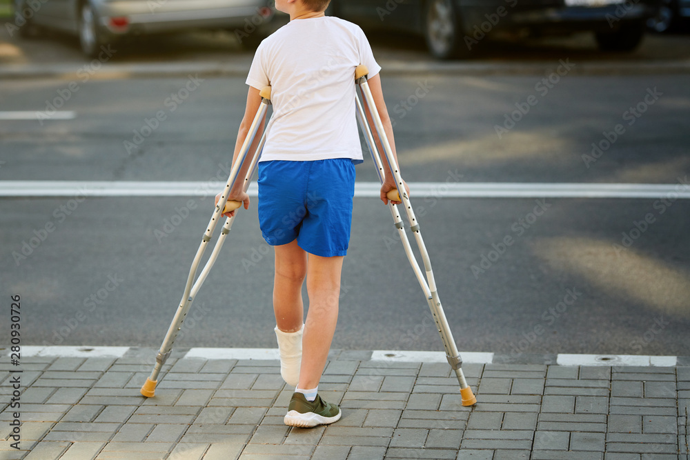 Young boy in orthopedic cast on crutches walking on the street near the road.  Child with a broken leg on crutches, ankle injury. Bone fracture and ankle  fracture in children Stock Photo