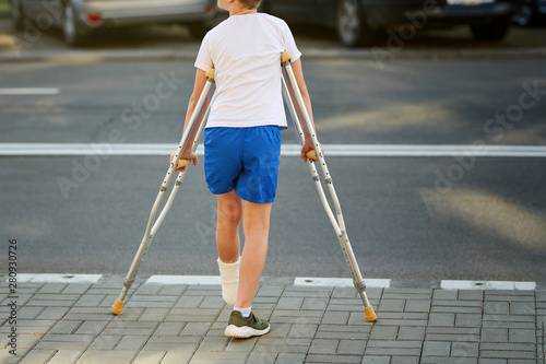 Carta da parati Young boy in orthopedic cast on crutches walking on the street near the road
