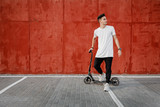 Young guy dressed in jeans and t-shirt stands with a scooter against a painted concrete wall on the summer day in the city.