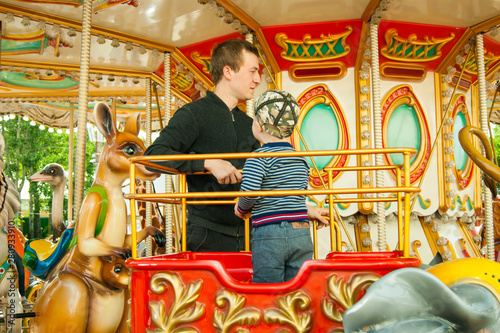 A three-year-old blond boy rides in a red open carriage on a merry-go-round, his dad is standing next to him. Fun, activity, impressions, emotions. © Наталия Пономарева