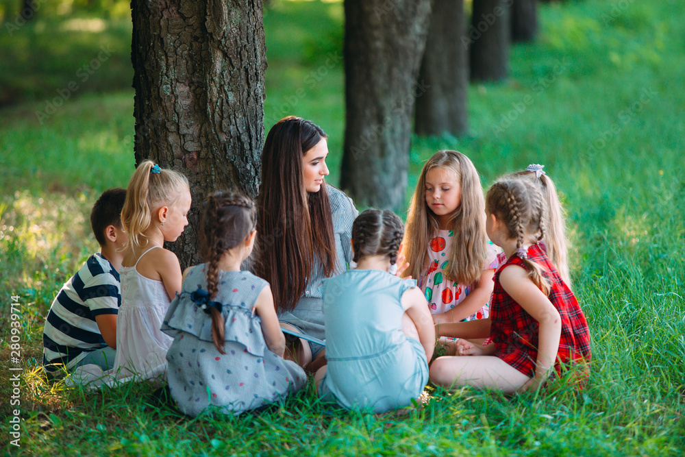 Children and education, young woman at work as educator reading book to boys and girls in park. Children and education, young woman at work as educator reading book to boys and girls in park.